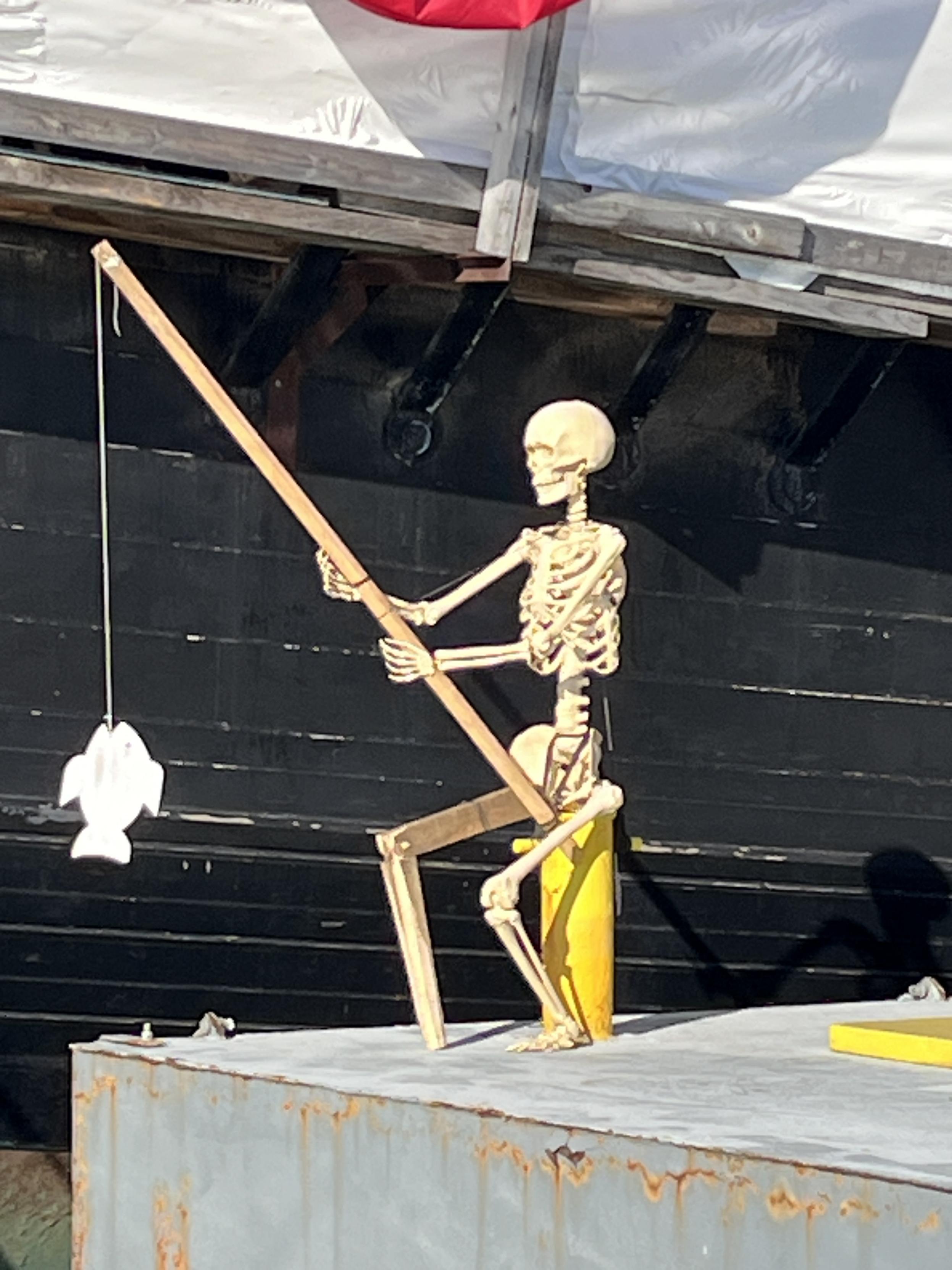 A skeleton seated on the dock next to the ship. The skeleton has a wooden leg and is "fishing," having hooked a fake fish.