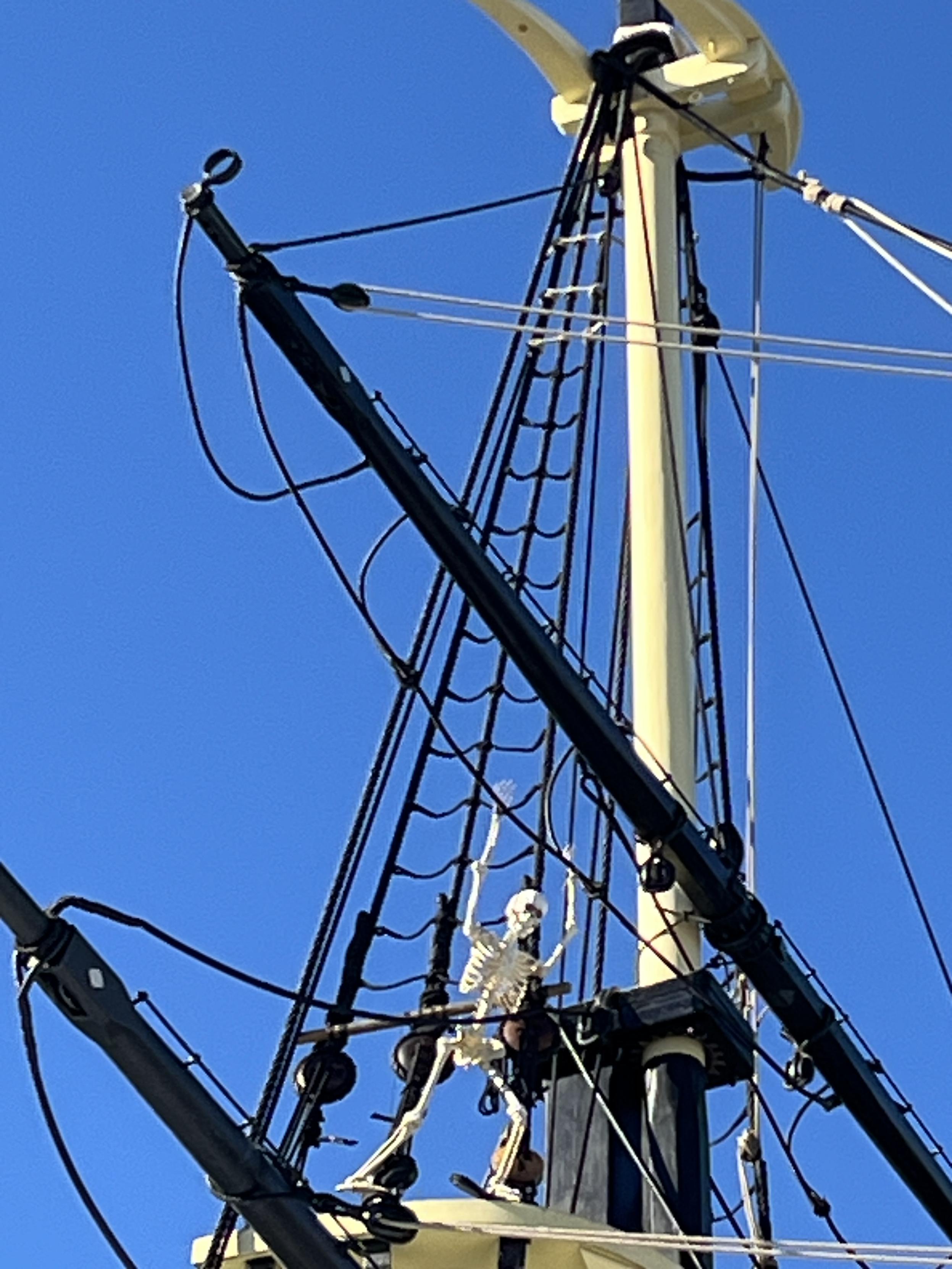 A skeleton is poised with arms raised in the ropes above a black spar on the ship. The masts are pale yellow. A skylarking skeleton, if you will.