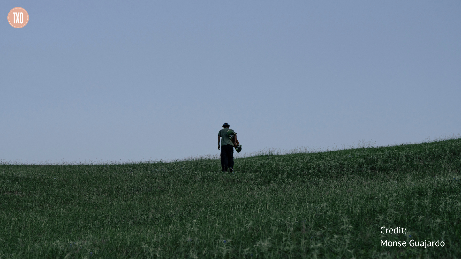 Robinson Gonzalez, seen from behind under a dusky sky, carrying his skateboard as he walks across a grassy, sloping landscape. Photo credit: Monse Guajardo