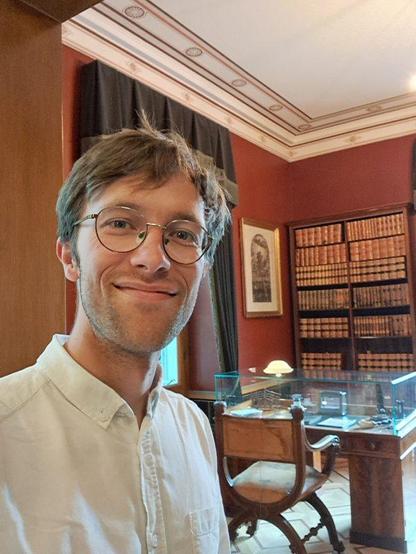 A smiling young researcher with glasses in a dark orange painted room in front of antique bookshelves and furniture