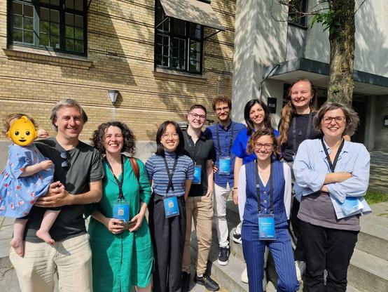 Nine researchers with blue conference badges, one holding a baby, smiling in the sun, in front of university buildings.
