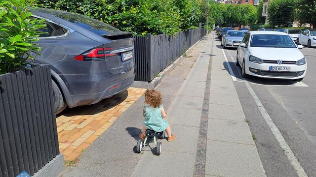 Small child in green dress riding tricycle on sidewalk, next to an SUV parking in driveway.