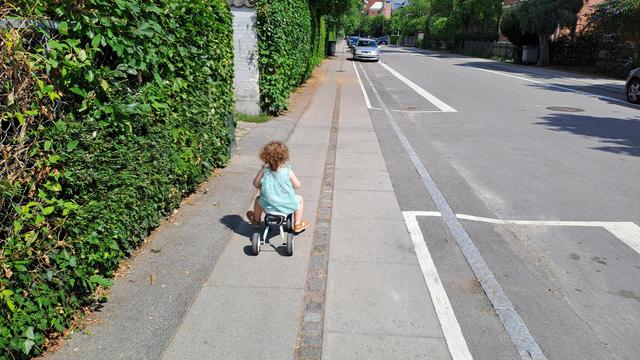 A small child in green dress riding a tricycle on sidewalk, with low-vision driveway approaching on the left from which an SUV could exit anytime.