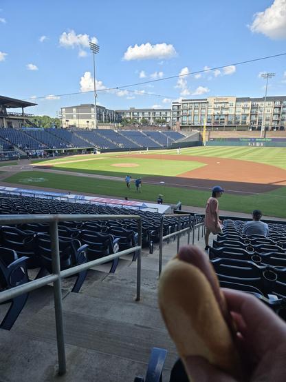a picture of a baseball field from the first base stands.  there are players warming up in the outfield on both sides and a hotdog being held up in the foreground.