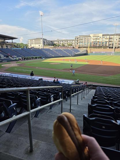 picture of a baseball field from the first base stands.  the grounds crew are getting the field ready.  a hot dog is visible in the foreground.