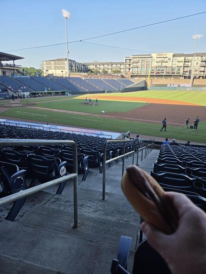 picture of a baseball field from the first base stands.  the grounds crew is making the field ready.  a hot dog is visible in the foreground.