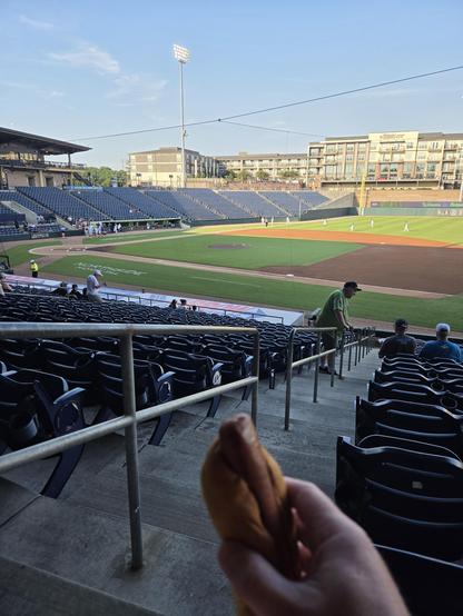 picture of a baseball field from the first base stands.  there are players working out and stretching in the outfield.  the infield is empty.  a hot dog is visible in the foreground.