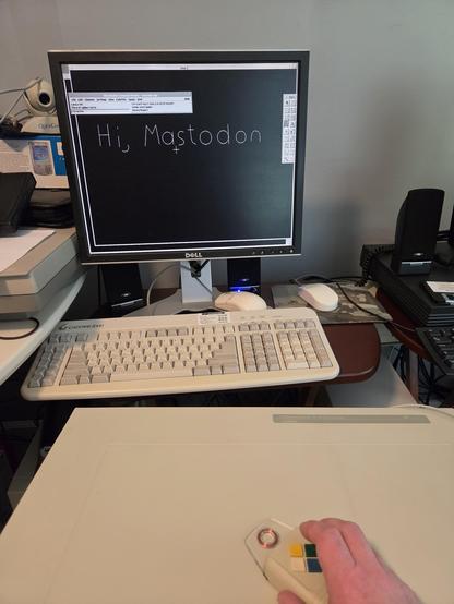picture of an LCD with a black screen and the  handmade words "Hi, Mastodon".  below that is a 1990's gateway 2000 keyboard and a large Summagraphics II digitizer board.  a hand is holding the 4-button pointer.  some other retro systems are visible to each side.