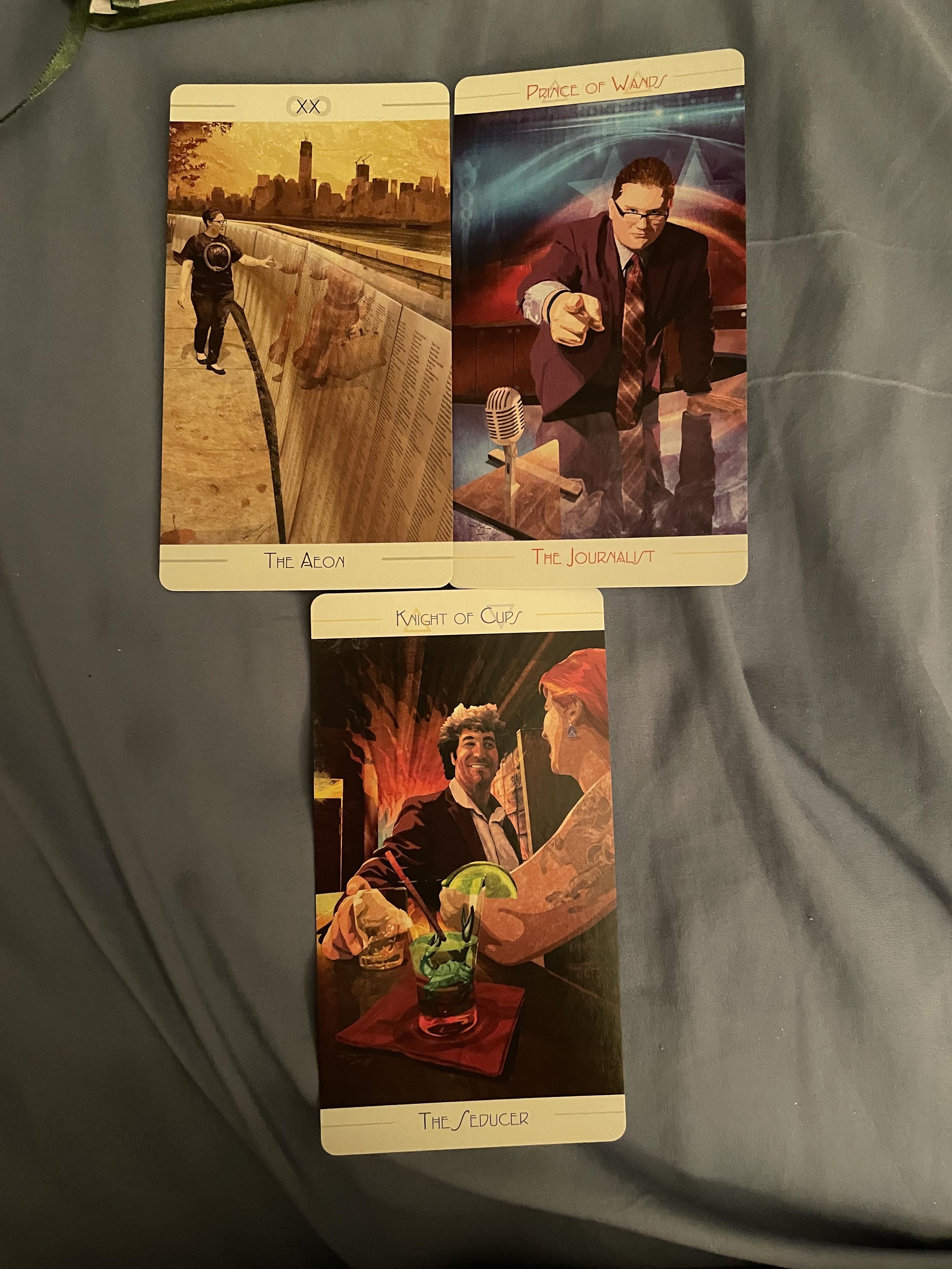 Two tarot cards side by side, and a third centred below them.

The first card is the Aeon, showing a woman walking by a memorial walk by a river, running her fingers over names.

The second is the Prince of Wands, subtitled the Journalist. It shows a man with short hair and glasses leading over a desk with a microphone, his finger extended accordingly at the audience. The background has the colours of the US flag.

The third card is the Knight of Cups, subtitled the Seducer, It shows a man with shaggy coiffed hair and wide-collared suit sitting at a bar chatting a woman up. There is a crab in her cocktail glass and a horse tattoo on her arm.