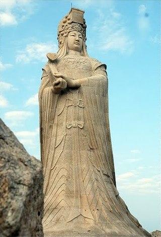 A statue of Mazu at Meizhou Mazu Temple on Fujian. She stands radiant in worked stone, her dress tied tight, her headdress massive and layered, her face calm and serene.
