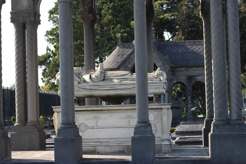 A grave at Glasnevin Cemetery, showing a man sleeping above an above ground crypt, surrounded by pillars.