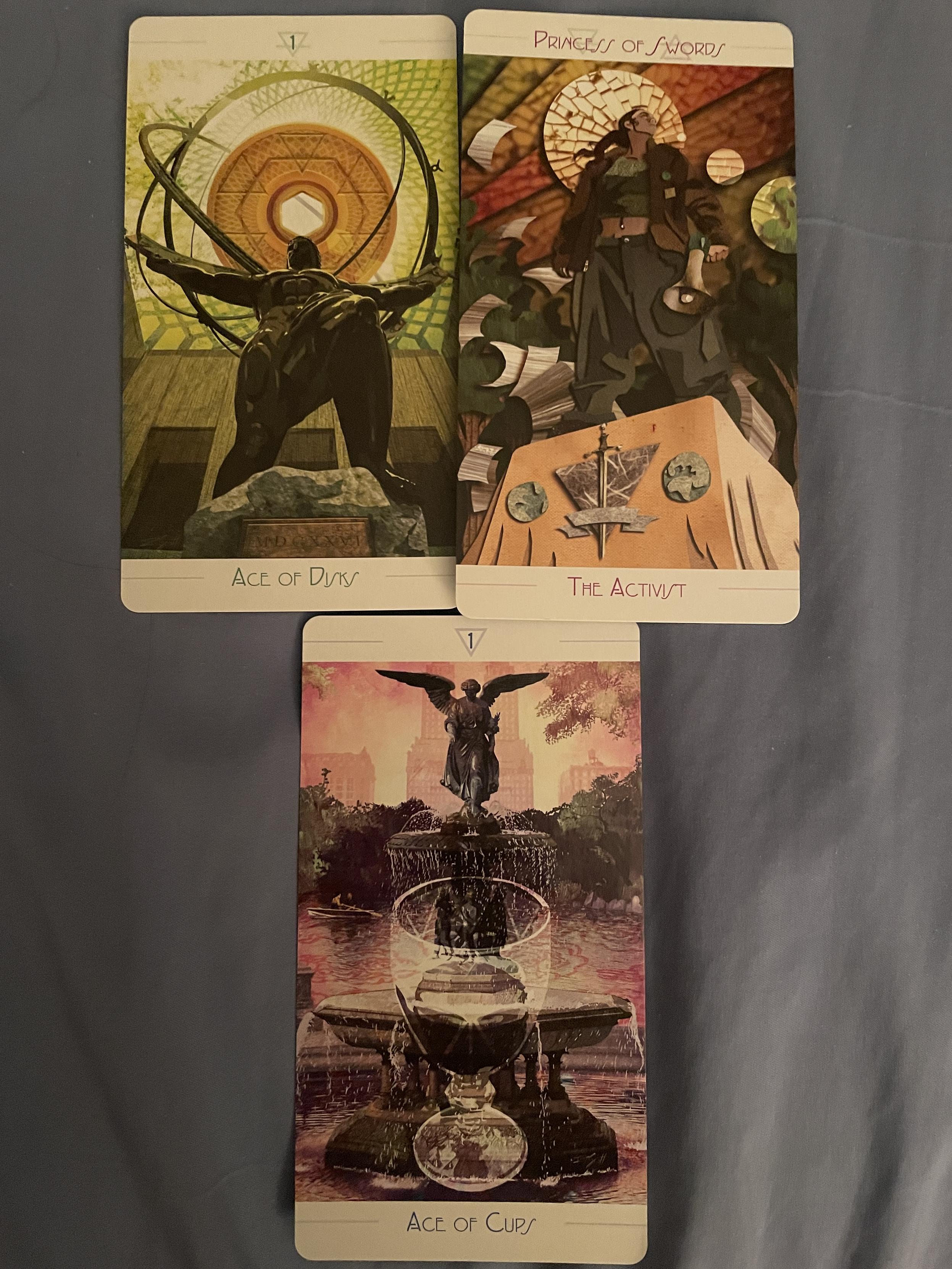 An inverted pyramid of three tarot cards. The top two are the Ace of Disks and Princess of Swords, the bottom one the Ace of Cups.

The Ace of Disks shows an Atlas statue cast in metal, where he's holding metal circles above his head. Behind him is a single Disk, modeled on a New York City subway token of a certain vintage. The Princess of Swords wears grey pants, a belly button ring, a crop top and a brown jacket. She is holding a megaphone, standing on a plinth with a single sword on it. Stacks of paper surround her, the top pages being blown away by the wind. The Ace of Cups shows a fountain with an angel on top, and a pond on which people are rowing behind the fountain. Behind the pond are trees, and in the distant background, skyscrapers.