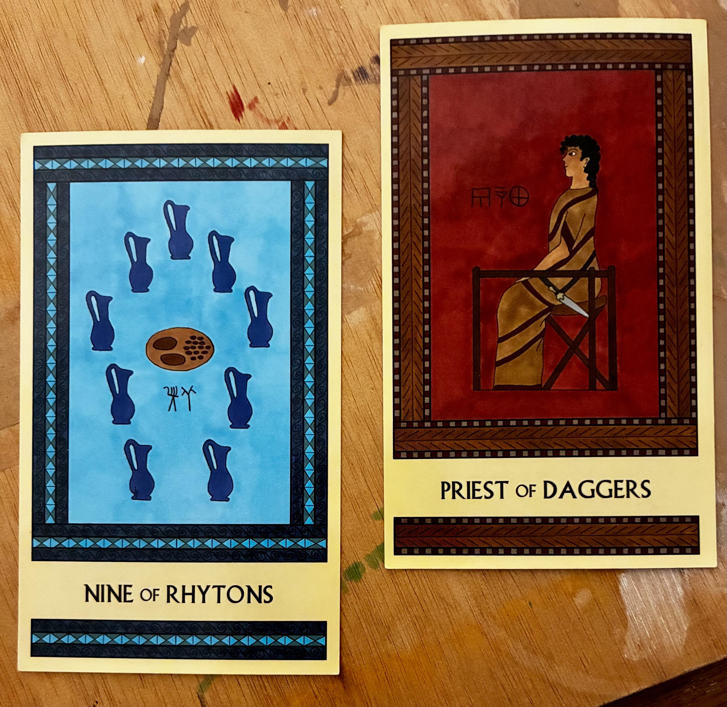 Two Minoan Tarot cards side by side on my ink-and-paint-splattered plywood art table. Both cards have intricate rectangular borders that are repeated in a strip at the bottom of the card, below the card's title. The card on the left, the Nine of Rhytons, is painted in shades of blue. It depicts nine royal blue pitchers arrayed in an oval around a platter of olives and loaves of bread. The card on the right, the Priest of Daggers, is painted in shades of deep red and brown. It depicts a Minoan man in a long robe. He is seated on a stool, facing left, with a dagger hanging from a belt slung around his hips.