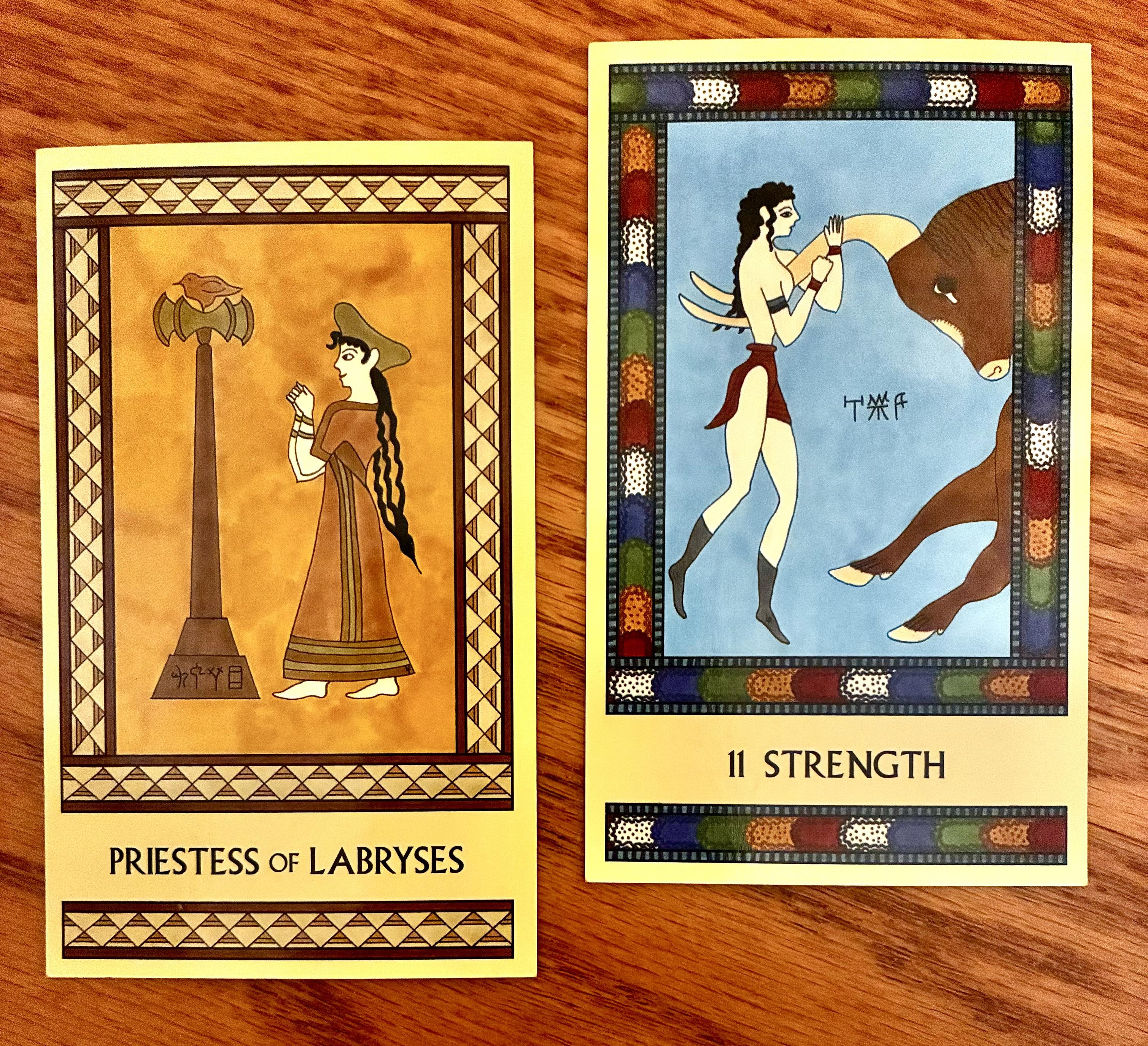 Two Minoan Tarot cards side by side on a golden brown wood surface. Both cards have intricate rectangular borders that are repeated at the bottom of the card, below the card's name. The card on the left, the Priestess of Labryses, is painted in shades of golden yellow, tan, and cream. It depicts a Minoan woman in a long robe and a large hat. She has long, wavy black hair that trails down her back almost to her knees. She is standing, facing left, with her arms raised in front of her and her hands clenched into fists. To her left is a tall double labrys in a stand, with a bird perched on top of it. The Strength card, on the right, has a bright multicolored border. It depicts a Minoan woman in a loincloth and boots, with long black hair. She is facing right, hanging from the long, curved horn of a brown bull that is charging in from the right. Only the head and forelegs of the bull are visible on the card.