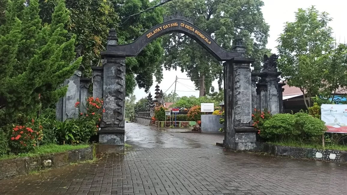 Bengkala's gate in modern Indonesia, a stone archway with Indonesian flare on both brick and concrete roads and the village beyond with high gardens and stone barriers to prevent trespassing. It is verdant, colorful, and very wet, as if after a summer rain.