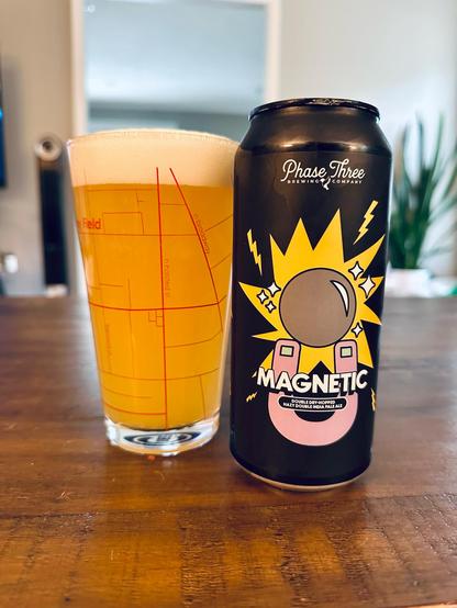 A pint glass full of beer next to a black can of Phase Three Magnetic beer