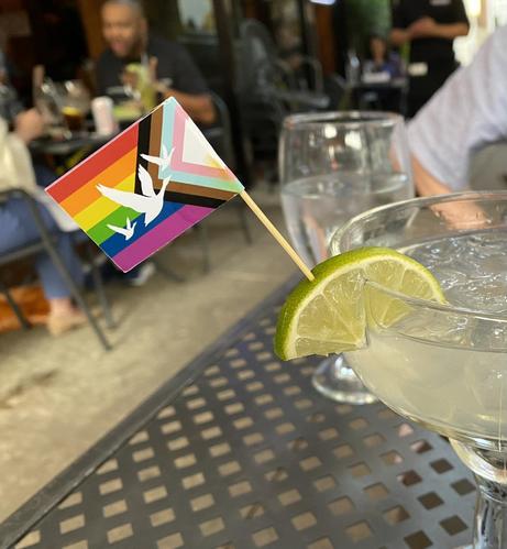 A large margarita glass extends off the right side of the photo, sitting on a metal outdoor restaurant table.  The drink is garnished with a lime slice, into which has been stuck a Pride flag featuring a rainbow, and then triangles of black, brown, light blue, pink, and white -- I think this is the Philly pride flag?  The loveliness of this small flag is undermined only slightly by the white geese flying across it, which look like WaWa logo business...