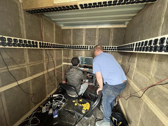 Two men are inside a small room baffled with sound dampening panels on the walls and surrounded by over 250 small speakers mounted on the sides and overhead. One of them sits at a computer while the other leans over his shoulder. There are wires and tools everywhere.