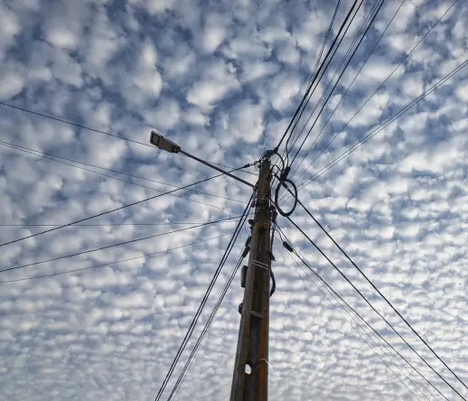Photo: Powerline pole with a street lamp attached and cables spreading in all directions. In the background, a blue sky filled with small clouds.