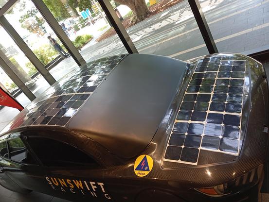 UNSW Solar Car. Black carbon fibre body. Body like a hatchback. Flat solar panels over the bonnet and roof. Windows blacked out with plastic shield. UNSWIFT Racing logo on door panel, and a sponsor badge near bottom left front window, in yellow.