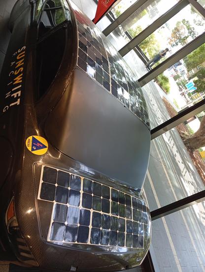 UNSW Solar Car. Black carbon fibre body. Body like a hatchback. Flat solar panels over the bonnet and roof. Windows blacked out with plastic shield. UNSWIFT Racing logo on door panel, and a sponsor badge near bottom left front window, in yellow.