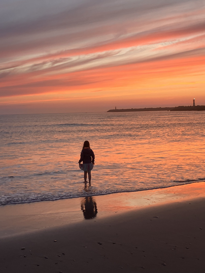 my daughter standing in the ocean in the sunset
the waves are at her ankles
the colours are orange, pink pale blue, various greys
foreground, bottom dark sand
then wet sand, part of the girl is reflected in the wet sand
then the water.  the girl is looking away, towards the sunset
there is a breakwater jutting out into the center from the right
the sky is bands of fire and greys and the light blue, looks a little like Jupiter.