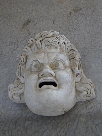 Eine weisse Maske mit grossen hervorquellenden Augen und einem nach unten gezogenem offenen Mund.
Wirre lockige Haare umrahmen das Gesicht.
