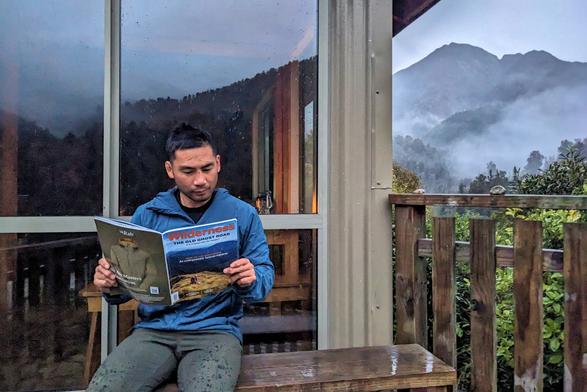 Me reading Wilderness Magazine about Old Ghost Road on a bench outside Specimen Point Hut (in the rain; my pants have some raindrops), with the large windows of the hut behind me and some mountains to the right. This picture got published in Wilderness Magazine...