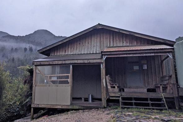 Specimen Point Hut is in the hills. It has a covered bike area to the left and stairs and a front door to the right.