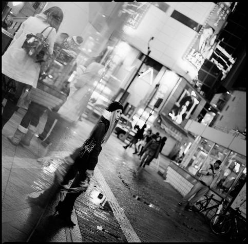 black and white photograph of and through a woman standing by a curved window sucking a lollipop.