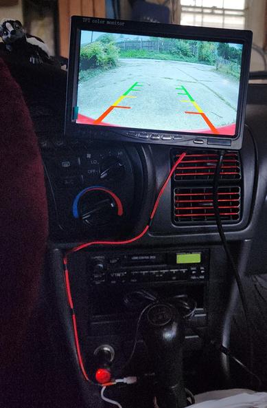 Black interior of a car with a backup camera screen floating at head height, beneath it the air louvers and power wire both in bright red, then the radio and the stick shift. You can also see a portion of the steering wheel covered in a dark red sheepskin cover.
The screen shows a cracked concrete driveway on a downslope with greenery on all sides, a chain link fence to the right, and a tall wooden fence on the other side of the street at the bottom of the drive.
