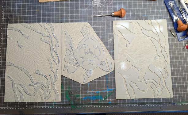 Three linocut printing plates next to each other. They show, from left to right: flowing strands of kelp that mostly leaves the center free, a fish with open mouth and huge eyes that fills the format's center and is staring right at the viewer, and the negative space that is not covered by either of the first - water.