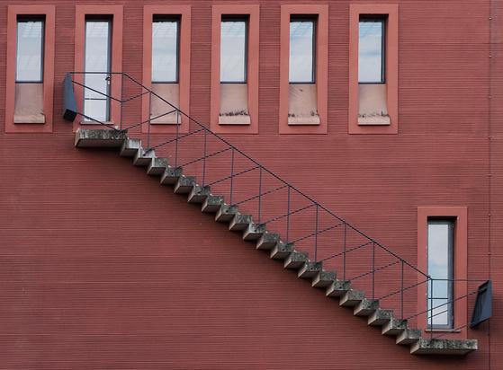 Rot verputzte Wand mit Fensterreihe oben und schmaler Tür rechts unten, verbunden durch eine Außentreppe.