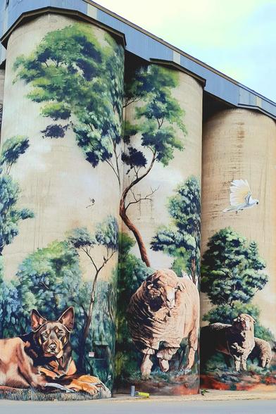 Three grain silos with a bush scene mural involving trees, sheep, and a kelpie.