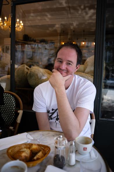 Une homme qui pose pour la caméra, son main sur son menton, assis a côté d’une table avec des tasses de café, et du pain.
