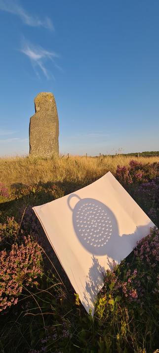 A modern version of an ancient standing stone on top of a heather clad moor, and in front of a summery blue sky. In the foreground, balanced in the heather, there is a piece of white paper. The shadow of a veg steaming pan is shown, with the solar eclipse curving the normally round holes into crescents.