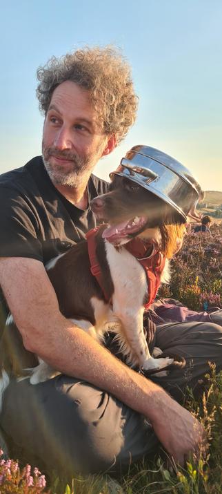 A white man with curly hair, holding a white and brown springer spaniel dog. The dog is wearing the veg steamer pan on her head. They might not the doing this eclipse thing right.l but they look beautiful in the low sun.