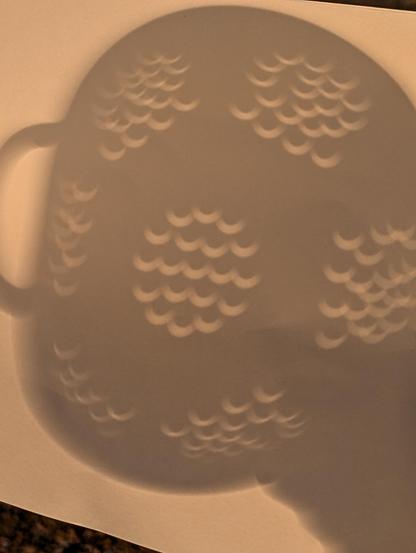 Partial solar eclipse seen through a colander.. basically a lot of crescent shaped holes in a shadow on a piece of card.