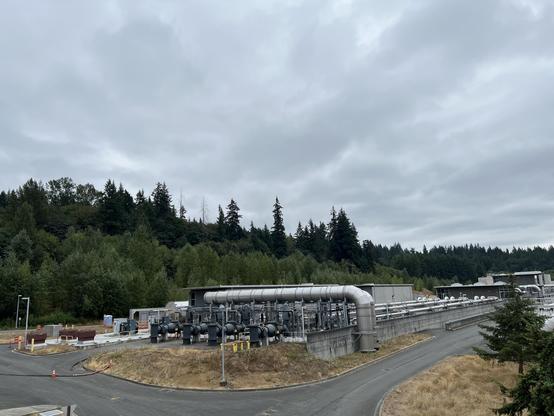 Picture of a very tree filled hillside and lots of the above ground buildings part of the Brightwater waster water processing facility.
