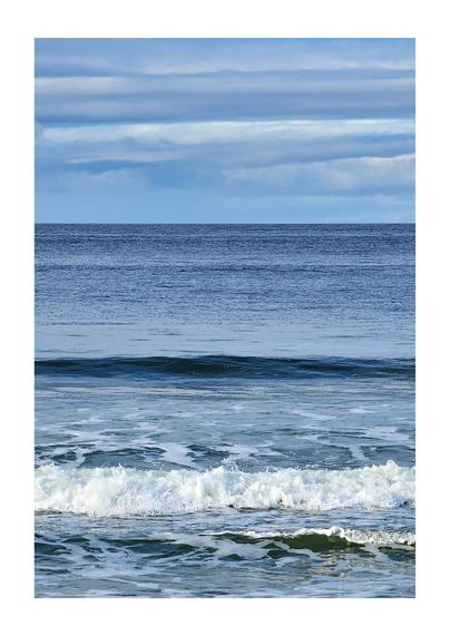 A portrait oriented photo of a view from a beach. The sky is a horizontal wash of white and light and dark blues, the horizon being about a third down the image, where the sea is a striped mix of dark and light blue lines until in the midground a dark blue horizontal band as a wave is about to break. In the foreground is a white bubbled horizontal band after another wave has broken.