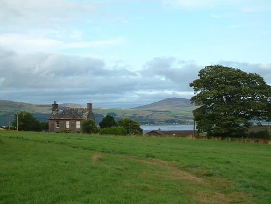A path curves through the foreground grass, a house to the left a tree to the right, between them the Solway Firth, in the distance green hills, clouds and blue shy.
