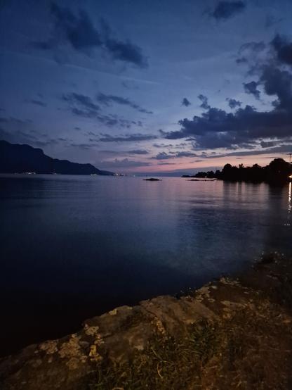 Lac Leman at night, August 2026
Still some red and pink to the right in the background. View in thr distance with the foot of an mountain to the left. The water is calm. Some clouds. Rocks right in front.