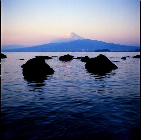 colour photograph of mt. fuji taken across a body of water (surge bay) in west Izu, japan. the peak of the mountain is clearly visible in the setting sun, a line of clouds stretches out below the peak. in the foreground dark shapes of rocks can be seen in the still water.