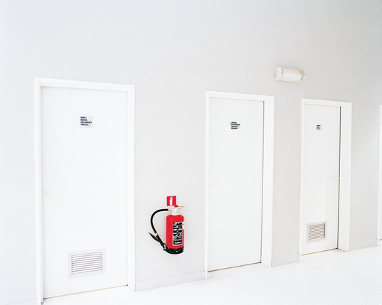 The described installation. The wall, floor and doors are stark white. The doors have signage in Dutch, French, English and German reading 'men, women and artists' respectively. Slightly distracting from the serenity is a bright red fire extinguisher.