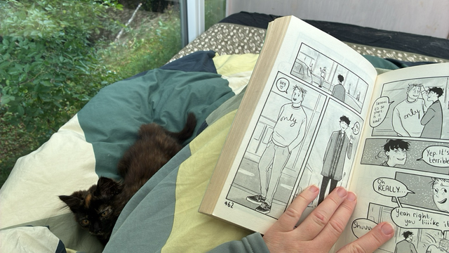 Photo of a white hand holding a graphic novel (Heartstopper) open. Next to the hand is a small tortoise kitten lying on the duvet
