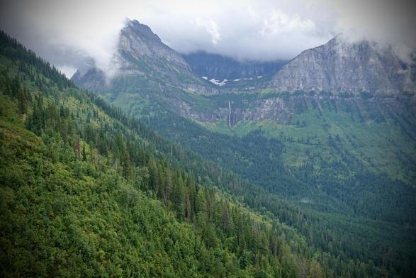 Image shows an expanse of two massive granitic peaks across two-thirds of the background, partially hidden in low-lying rain clouds and mist. Bare rock and sparse evergreen trees scale the front of the mountains, with a narrow line of snow. From the left, a sharp diagonal peak covered in trees reaches across the view to the right bottom frame.  (Glacier NP)