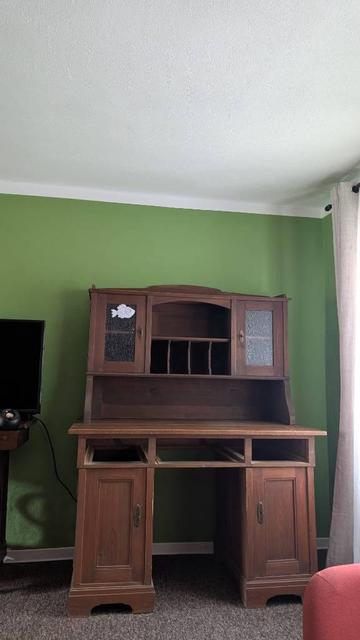 Eine grüne Zimmerwand und eine weiße Decke, davor steht ein antiker Schreibtisch in hellbraunem Holz mit einem massiven Unterteil und einem Aufsatz mit einem mittleren offenen Fach mit Unterteilungen und zwei Fächer mit Glastüren. Darunter weiter offener Raum über der Schreibfläche