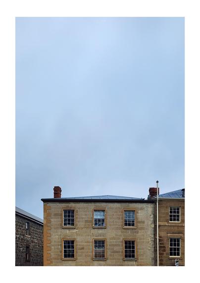 A portrait oriented photo of a gloomy, cloudy sky that takes up the top two-thirds of the image, with the lower third being the top two floors of multiple multistorey nineteenth century sandstone buildings with white framed windows and brick chimneys on low grey corrugated iron roofs.