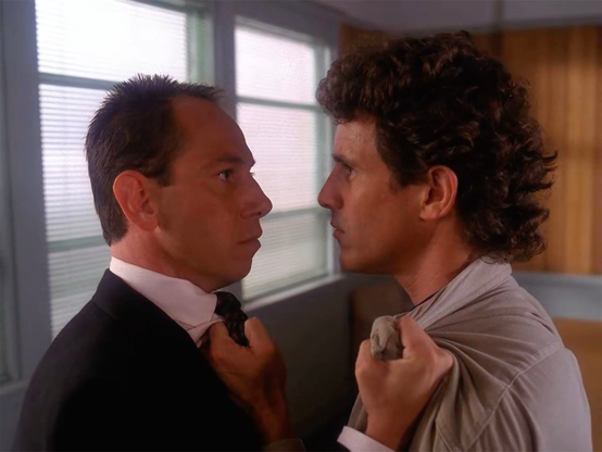 Special Agent Albert Rosenfield, played by Miguel Ferrer, stands on the left in a dark suit and white shirt. Sheriff Harry Truman, played by Michael Ontkean, is on the right, gripping Albert’s lapels with both hands and pulling him close. Their faces are only inches apart as they stare directly at one another, Albert appearing rigid and controlled while Truman looks openly angry and confrontational. Behind them is a brightly lit office with large windows covered by blinds and wood-paneled walls. The tight framing emphasizes the physical threat and comic intensity of the confrontation.