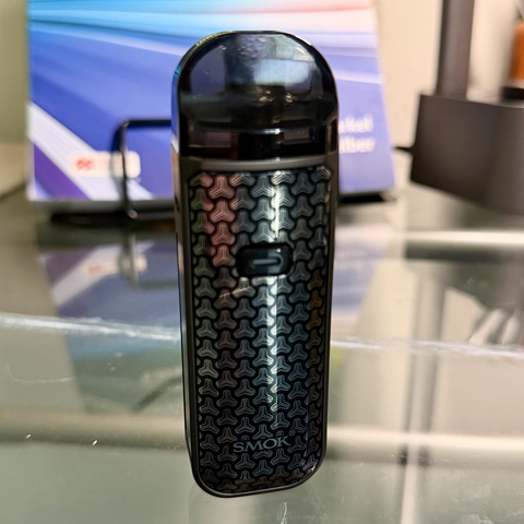 A plastic vaporizer device on a glass desk top. 
It is oblong and rectangular, rather like a two inch plinth. It is black and silver with a triangle shape pattern printed on it. A button is near the top. Near the bottom, the letters SMOK signify the manufacturer.